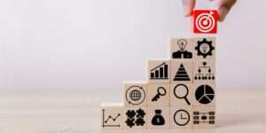 A hand placing a red wooden block with a bullseye target icon at the top of a rising staircase made of wooden blocks featuring business strategy icons.