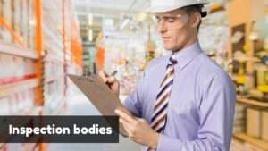 A male inspector wearing a white hard hat, safety glasses, a light purple button-down shirt, and a striped tie, holding a clipboard and writing with a pen inside a large warehouse aisle with tall shelves. A black text banner in the lower left corner reads "Inspection bodies".