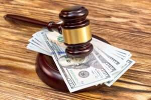 A wooden judge's gavel resting on a stack of United States one hundred dollar bills on a wooden surface.